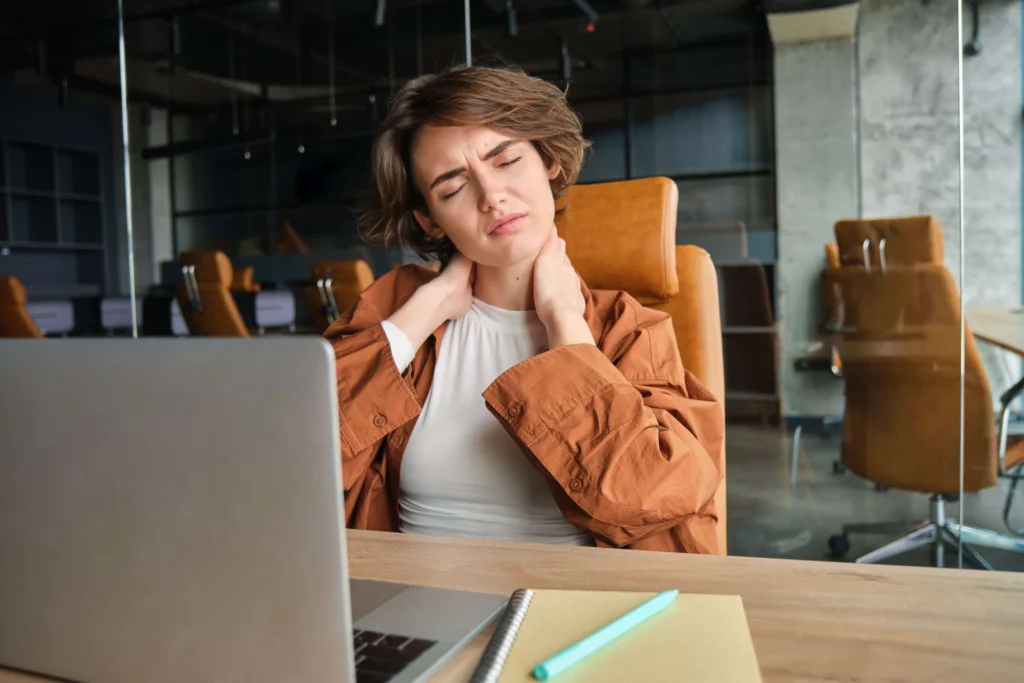 Young woman experiencing neck pain while sitting at a desk, holding her neck in discomfort and seeking a second opinion for her condition.