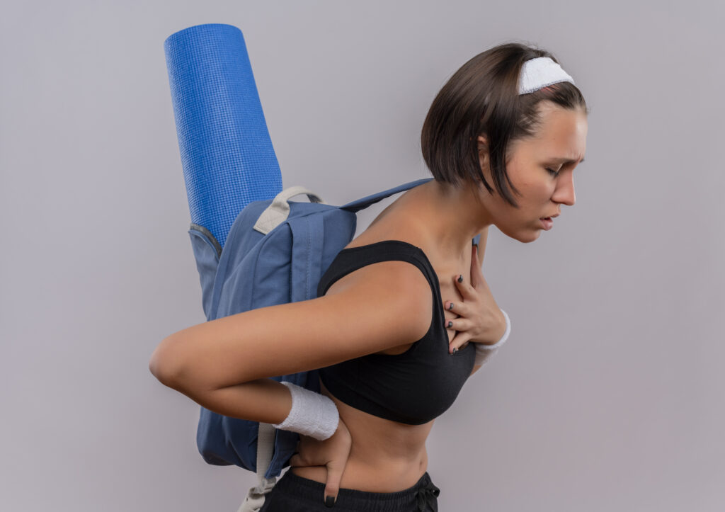 Young woman with back pain looking tired while carrying a yoga mat; exercises for brachial plexus injury recovery can help alleviate discomfort.