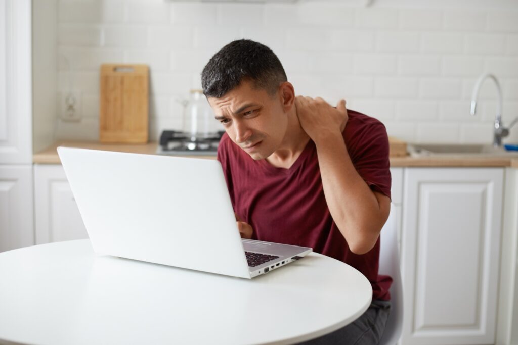 A man sitting at a kitchen table, rubbing his neck due to discomfort, which may be a sign of neck pain requiring attention from a spinal cord specialist.